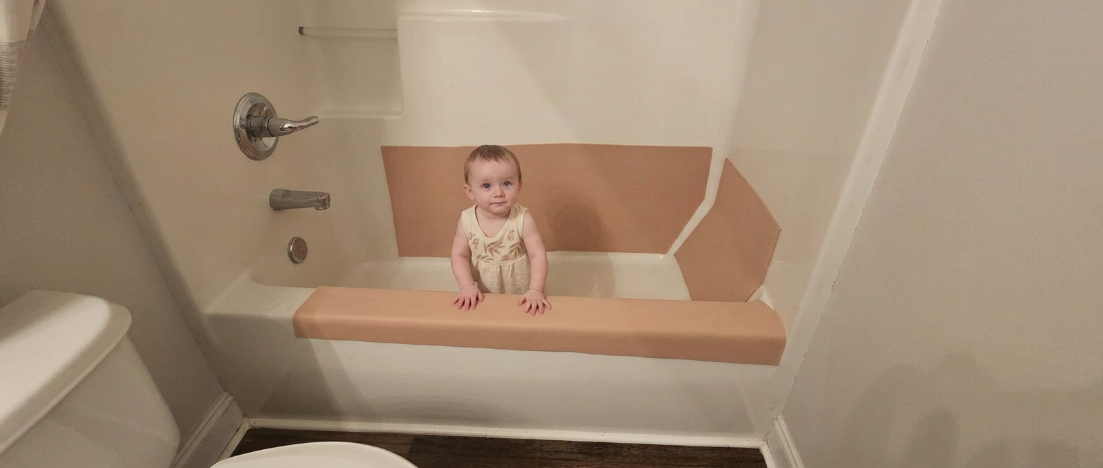 A baby standing in a bathtub with tan cushioning panels covering the back wall, side wall, and front tub edge.